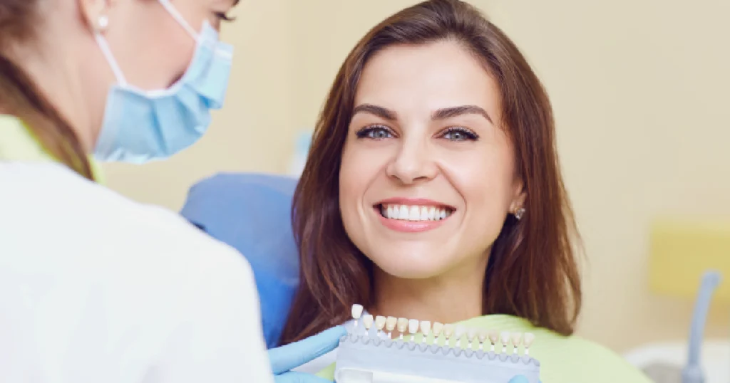 Smiling woman in a dental chair holding a tooth shade guide, with a masked dental professional assisting her during a KöR teeth whitening consultation or procedure in West Linn, OR